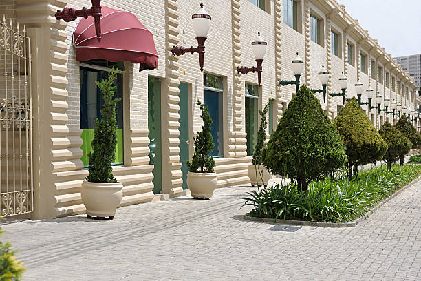 Charming retail shopping district with light brick building, red awnings, vintage wall lamps, stone walkway lined with large potted plants, trimmed hedges, and decorative flower beds