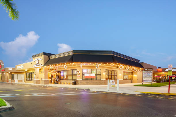 Commercial retail center building with warm yellow exterior walls, black awnings, decorative string lights, clean paved parking lot, palm trees, clear blue sky with white clouds — after professional stump grinding and landscaping services