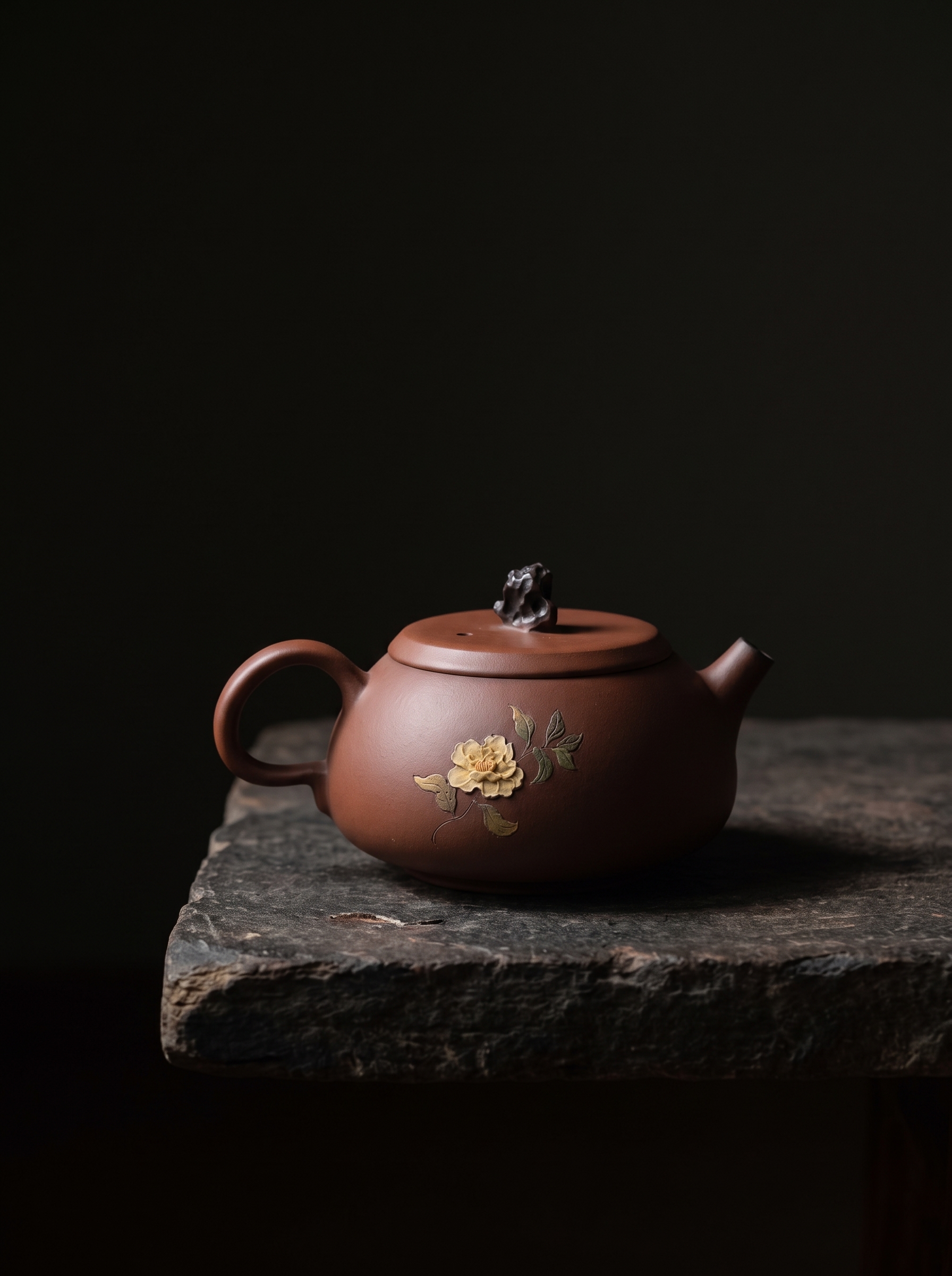 A brown-red Jianshui Zitao teapot inlaid with yellow flowers and green leaves, resting on a rough stone slab against a black background