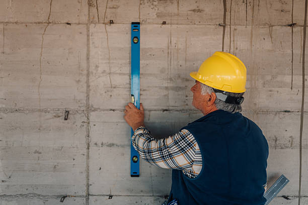 Construction worker in yellow hard hat checking poured concrete retaining wall with blue level during quality inspection to ensure wall plumbness and structural accuracy