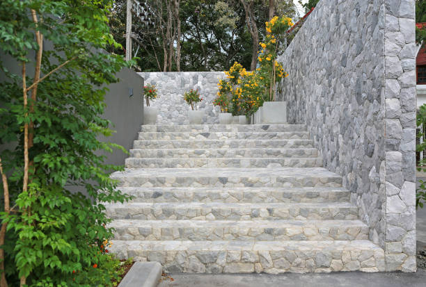 Natural stone retaining wall with stone steps flanked by lush greenery including trees and yellow flowering shrubs, creating a peaceful garden terrace landscape