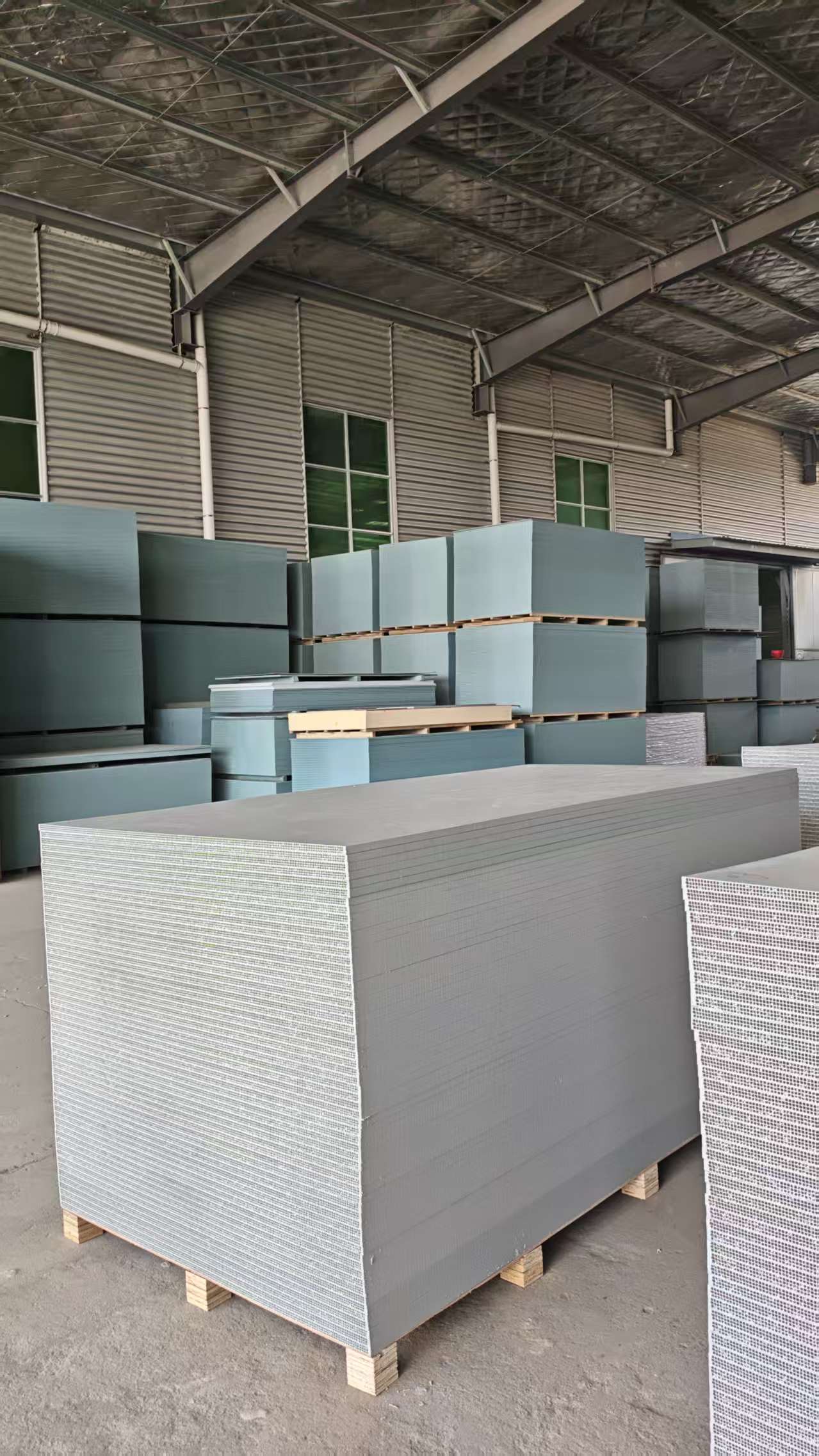 PP hollow plastic formwork panels in gray and blue-green stacked on wooden pallets in spacious well-lit warehouse with corrugated metal walls and windows
