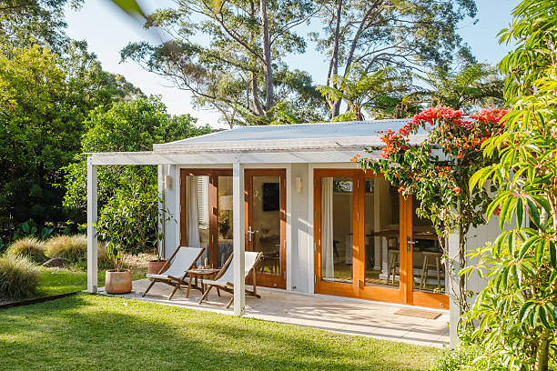 Complete outdoor living space with garden cabin featuring light colored walls wooden doors and windows wooden lounge chairs on well-maintained lawn surrounded by lush green trees flowers and plants creating a peaceful country style backyard retreat