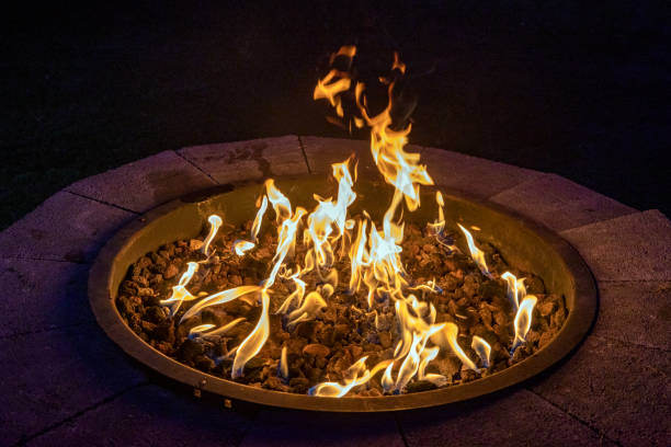 Professional gas fire pit installation showing circular metal fire bowl burning bright flames at night surrounded by decorative gravel and stone brick ring border in a dark outdoor backyard patio setting