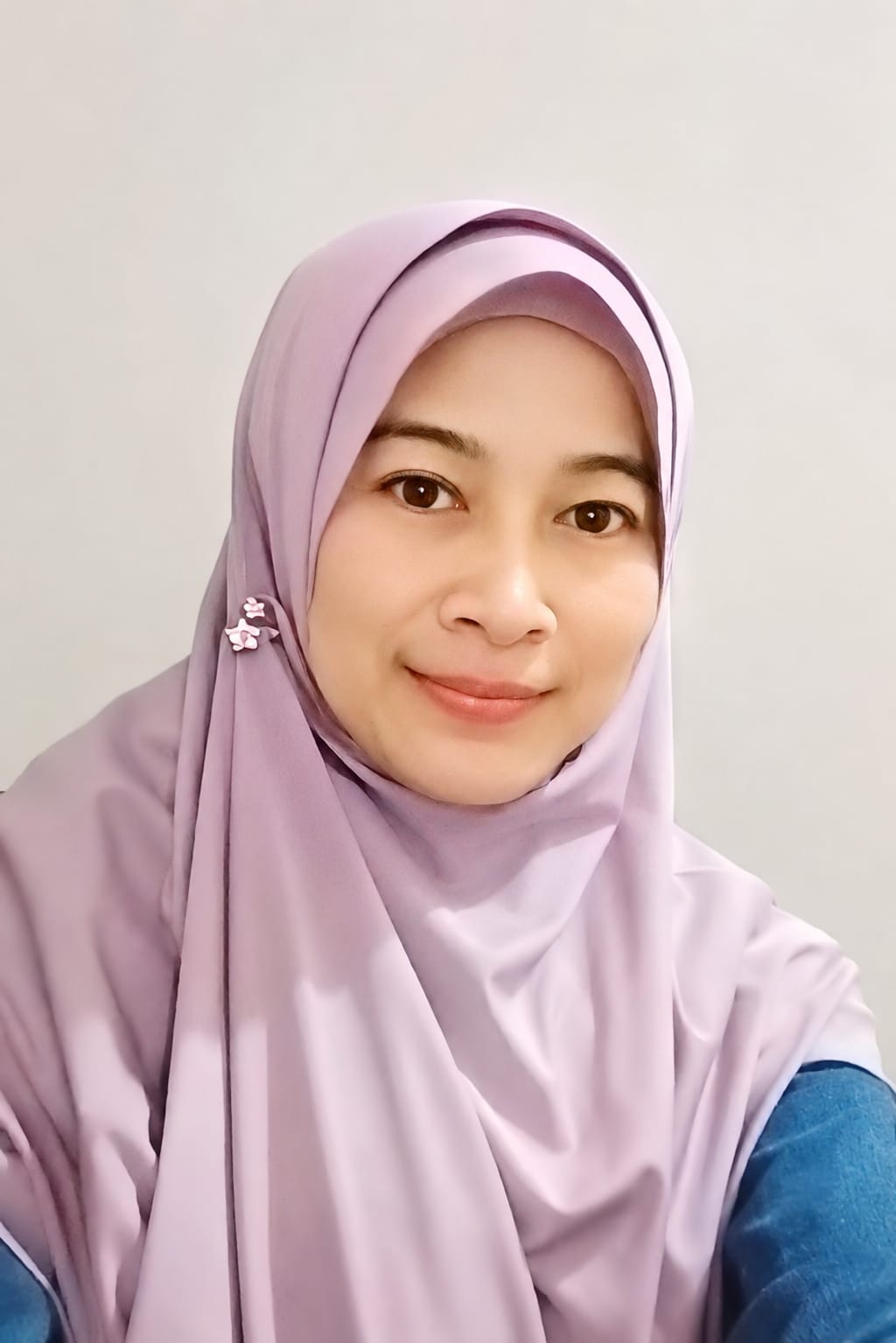 Potret Kak Mina Hanifatul Khairiyyah mengenakan hijab ungu muda dengan hiasan dan pakaian biru di depan dinding sederhana yang hangat.