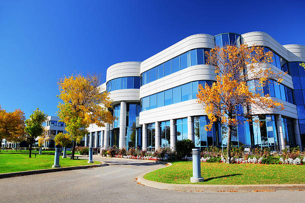 Modern corporate office park with glass and white architecture, manicured green lawn, walking paths, flower beds, and golden autumn trees under clear blue sky