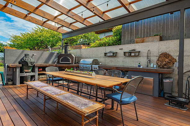Custom cedar pergola with outdoor kitchen featuring wooden deck, dining table, brick oven, grill, and stainless steel prep counter surrounded by green trees