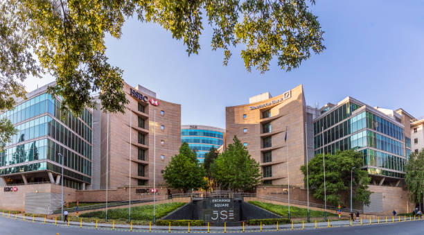Modern commercial office park complex with glass curtain wall and stone facade architecture, landscaped grounds with trees and green areas, 5S designated zone in foreground, clean roadways with railings, under clear blue sky