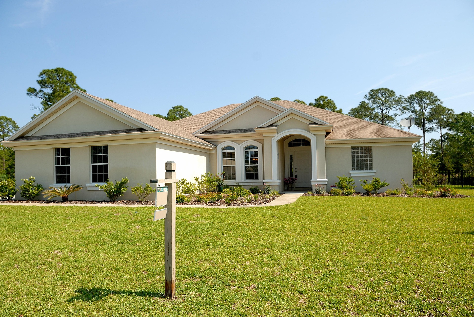 Single-family suburban home with light exterior and brown roof surrounded by evenly green lawn and healthy shrubs, demonstrating uniform sprinkler coverage from underground irrigation system on clear sunny day with blue sky