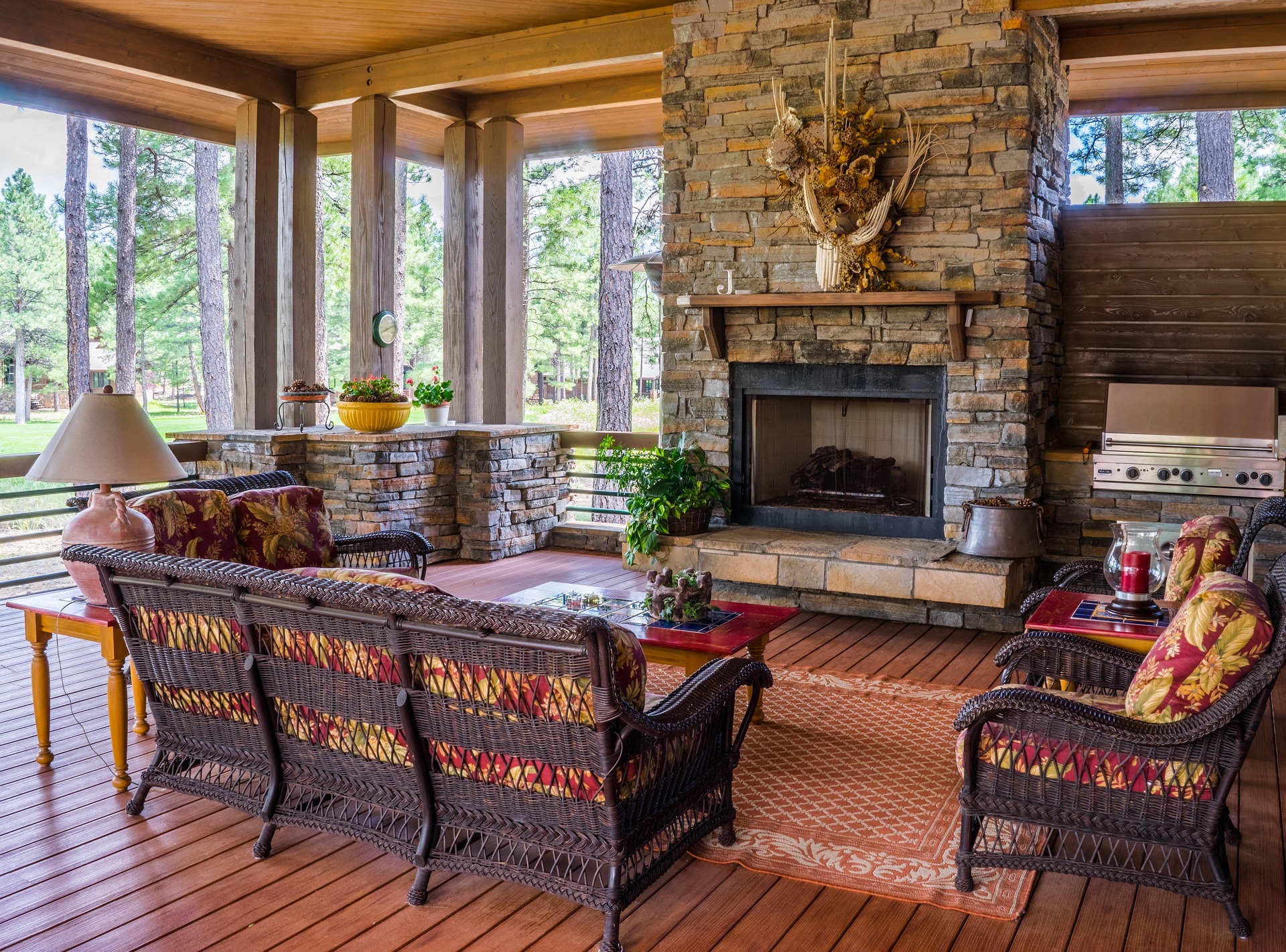 Custom outdoor kitchen installation in a forest setting with stone built-in fireplace rattan sofa with patterned cushions wooden dining table and chairs outdoor grill area surrounded by lush green trees creating a natural comfortable backyard living space