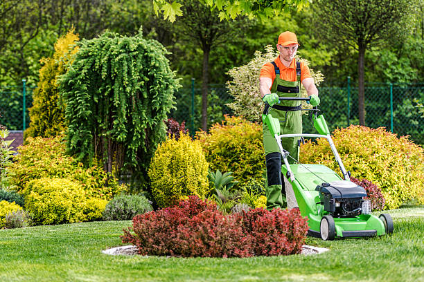 Worker in orange uniform mowing lawn with green mower near colorful garden shrubs