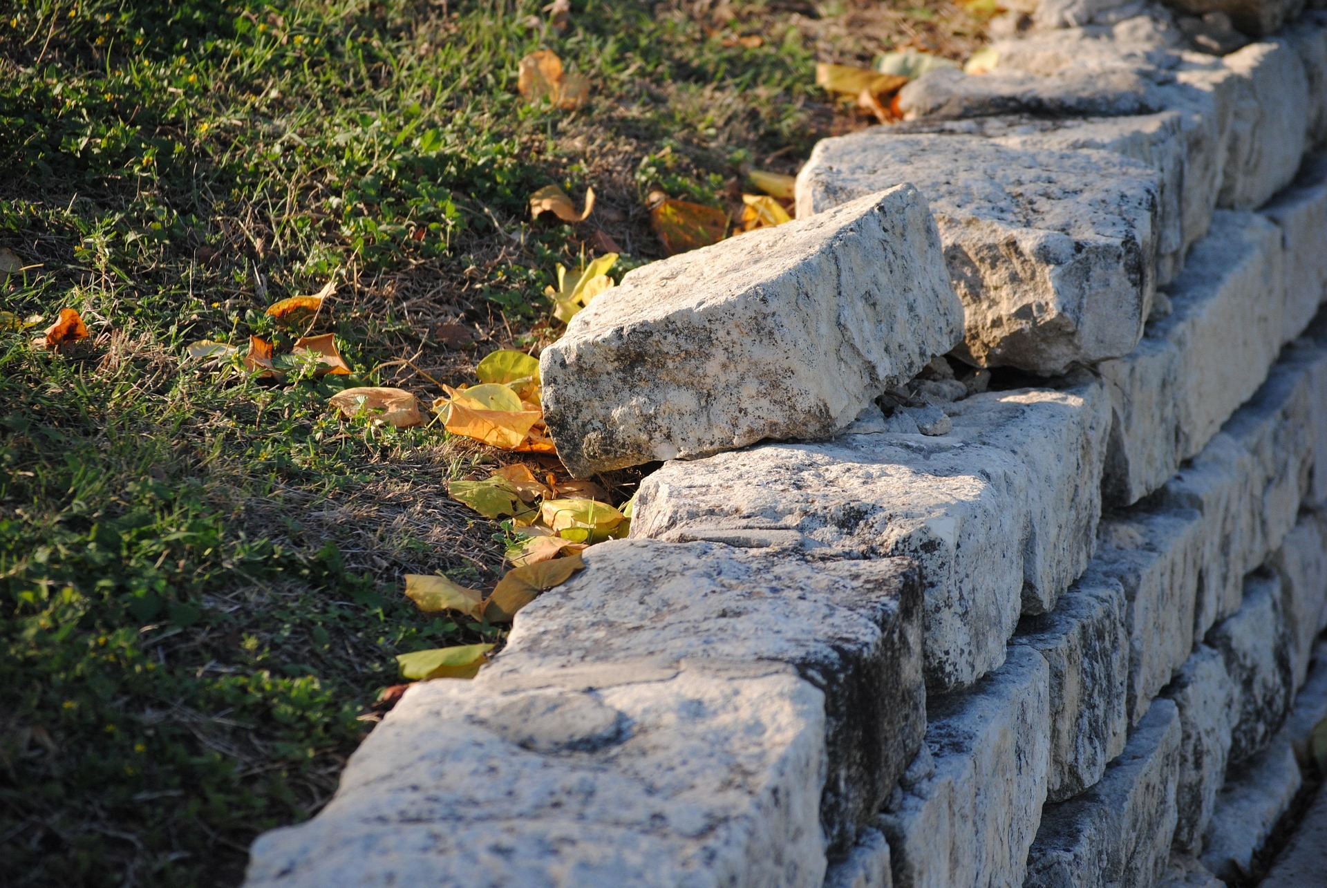 Weathered natural stone retaining wall with aged texture alongside grass lawn with autumn leaves, showcasing durable backyard terrace construction by Trusted Lawn Services