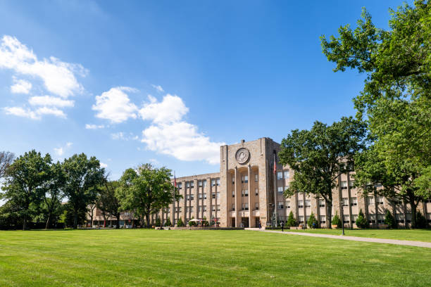 Educational campus with stately stone clock tower building surrounded by expansive green lawn, mature shade trees, and open blue sky with white clouds