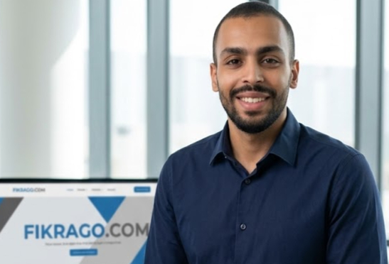 Ayoub Zinani, creator of Fikrago, standing in a bright office with Fikrago.com displayed on screen