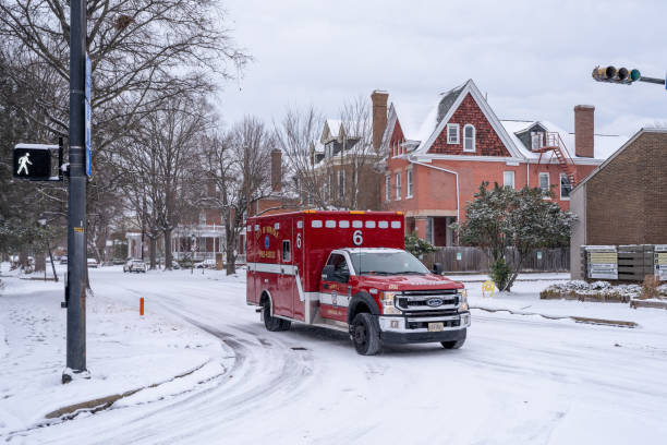 Red ambulance driving on snow-covered residential street with snow-buried houses and bare trees during winter blizzard conditions