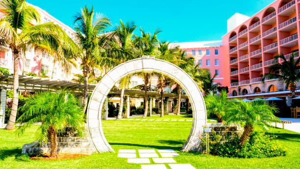 Tropical resort courtyard with stone archway surrounded by palm trees and lush green Bermuda sod lawn with pink hotel building in background on clear sunny day