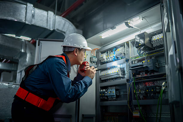 Industrial facility worker in white hard hat, blue uniform, and red safety harness inspecting electrical equipment cabinet with pipes and lighting in a warehouse factory environment