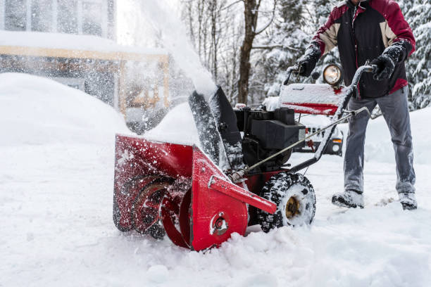 Worker operating red snow blower to clear deep snow from residential property with snow-covered trees and buildings in winter
